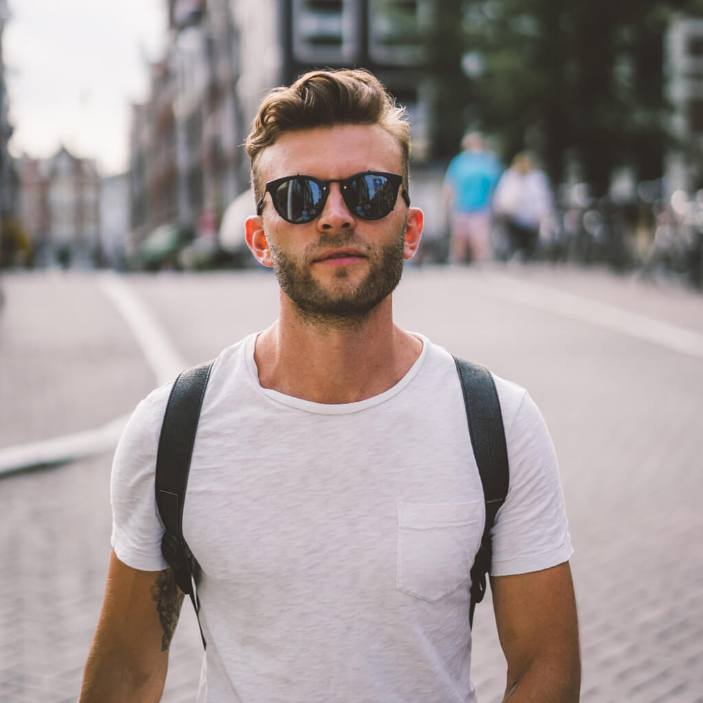 A stylish man in sunglasses walking on a city street, wearing a white t-shirt and backpack.