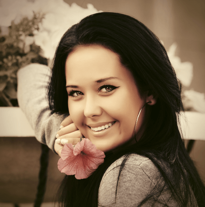 Smiling woman with black hair holding a flower, surrounded by nature and vibrant blooms.