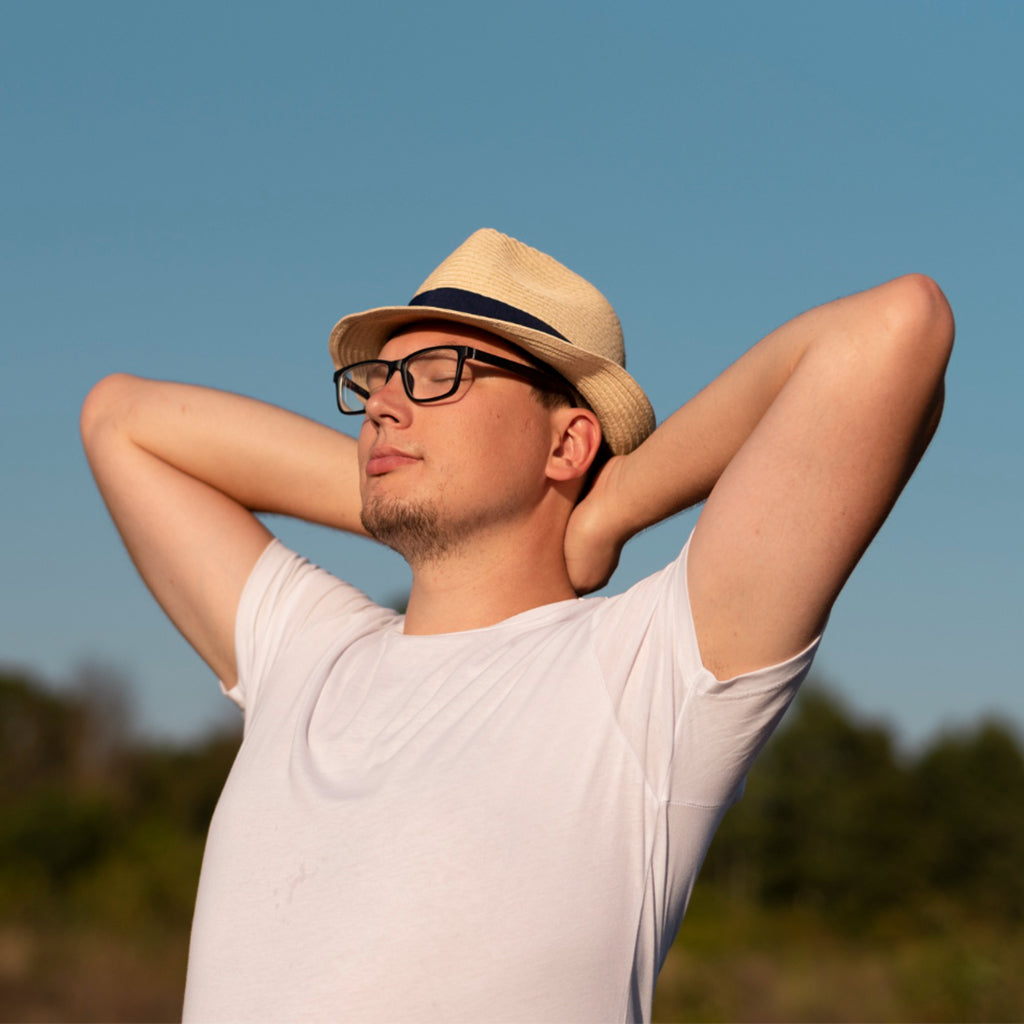 Man wearing a straw hat and glasses, relaxing outdoors with arms behind his head against a clear blue sky.