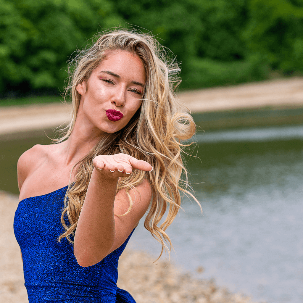 Model in a blue outfit blowing a kiss by a lakeside, surrounded by greenery.