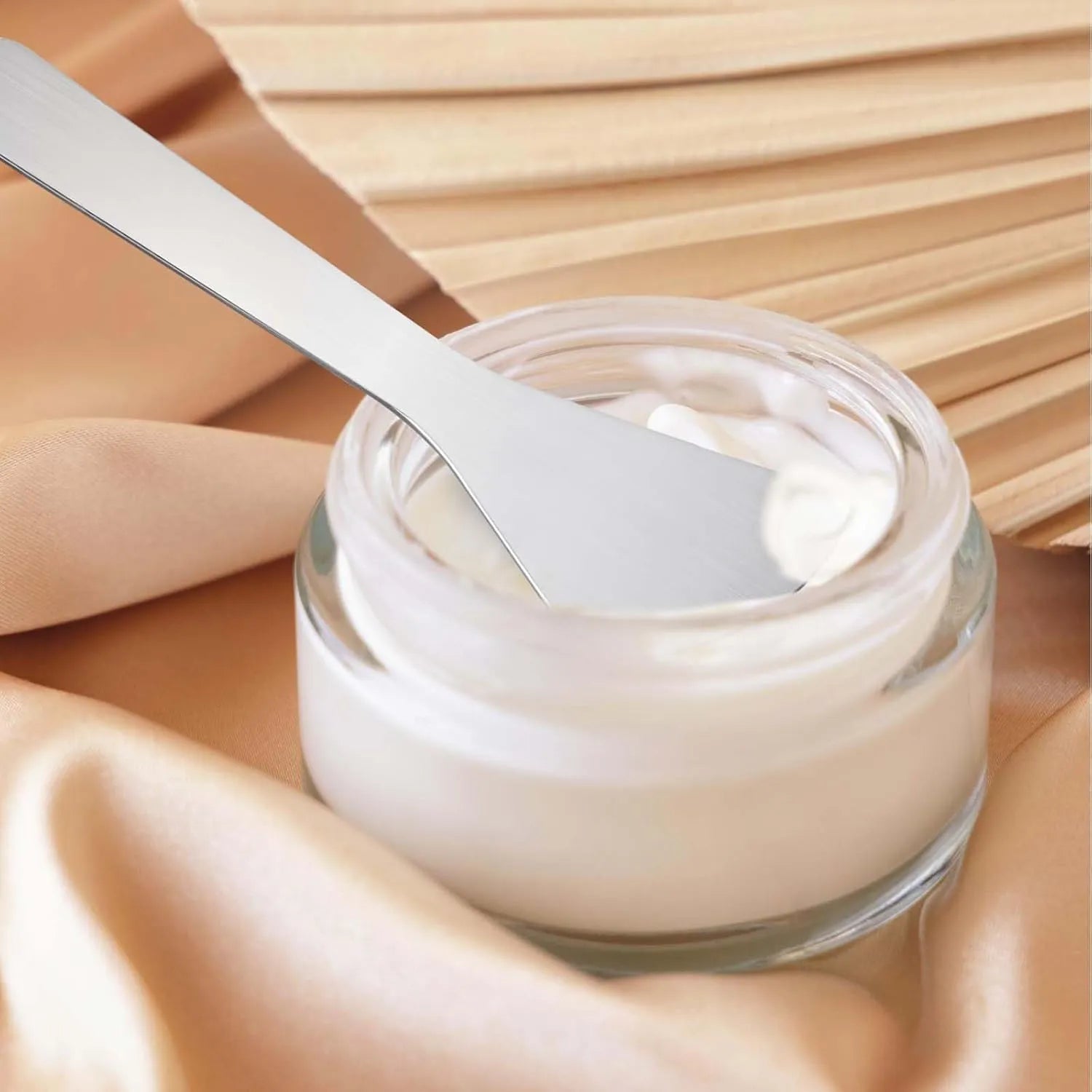 Stainless Steel Cosmetic Skincare Spatula Set with cream in a glass jar, on soft fabric background.