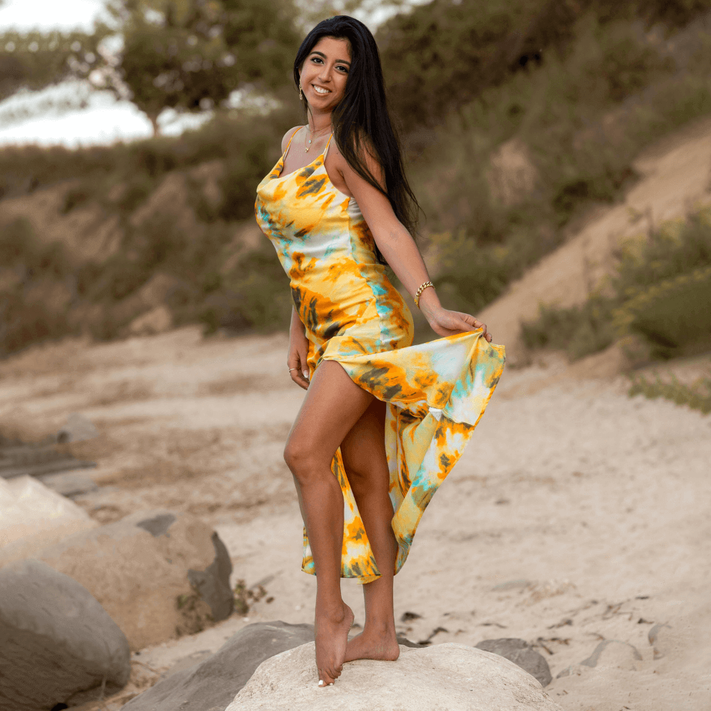 A woman wearing a colorful yellow and blue dress, smiling and posing on a beach, enjoying a sunny day.